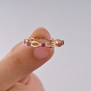 May include: A rose gold band ring featuring a repeating infinity-style openwork pattern interspersed with small, round, vibrant pinkish-red gemstones set at regular intervals along the entire circumference of the metal band.