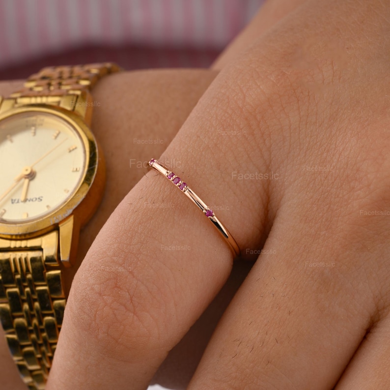 May include: A thin gold ring featuring three small, round, vibrant pink gemstones set along the band. The ring is worn on a finger next to a gold-toned analogue wristwatch with a metallic link strap and a round face with gold hands and markers.