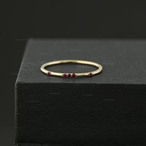 May include: A thin gold band ring featuring five small, round, deep red rubies set along the top half of the band. The ring has a smooth, polished finish and a delicate, minimalist profile, showcasing a simple design with evenly spaced gemstones.
