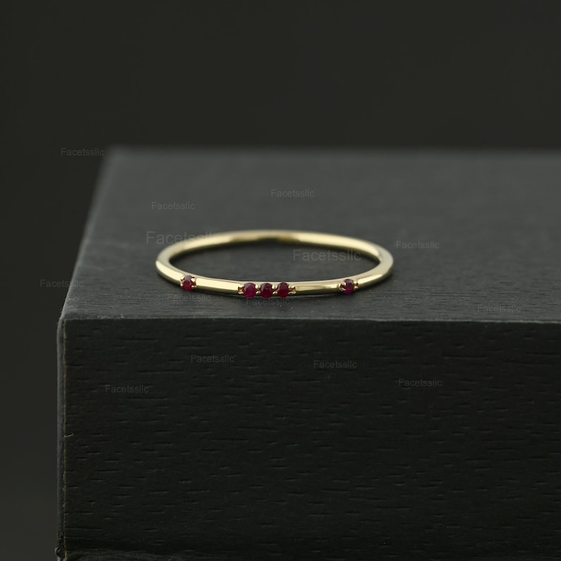 May include: A thin gold band ring featuring five small, round, deep red rubies set along the top half of the band. The ring has a smooth, polished finish and a delicate, minimalist profile, showcasing a simple design with evenly spaced gemstones.