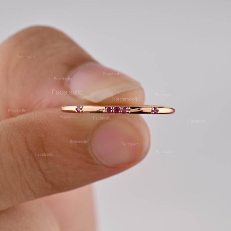May include: A thin rose gold band ring featuring a horizontal row of five small, round, deep pink rubies set into the metal. The ring has a smooth, polished finish and a minimalist design, showcasing the vibrant red gemstones against the metal.