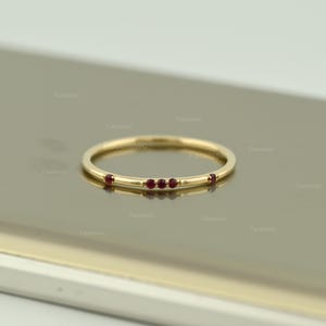 May include: A thin yellow gold band ring featuring five small, round, deep red rubies set along the top. The stones are arranged with a cluster of three in the centre and one single ruby on each side, creating a delicate and minimalist jewellery piece.