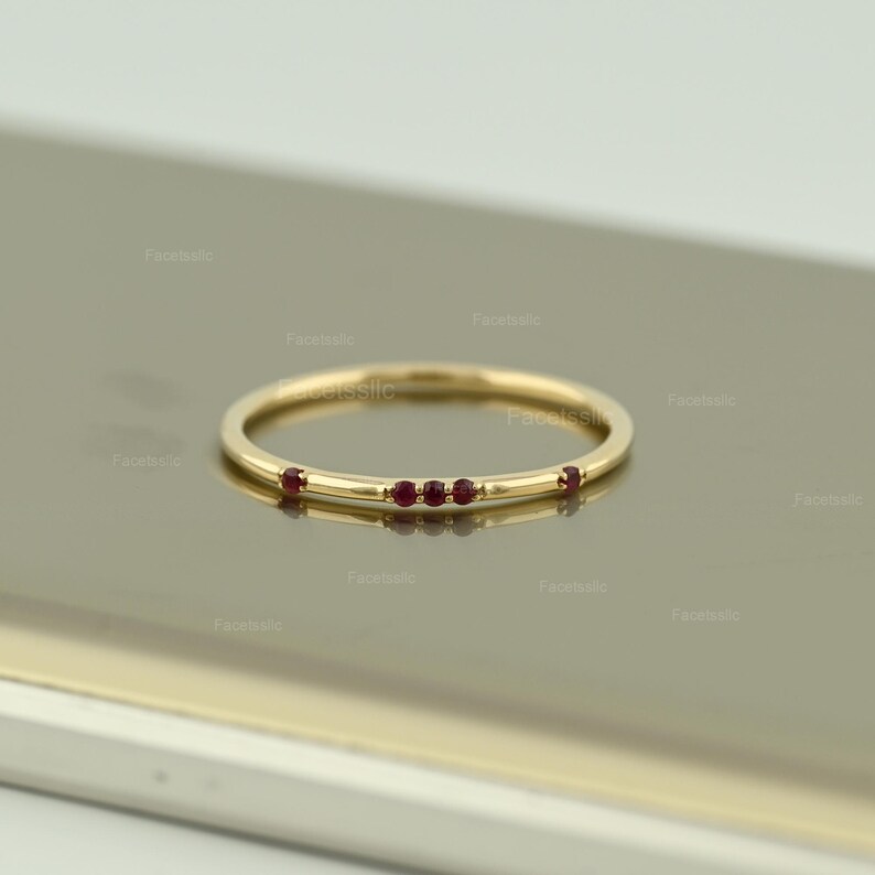 May include: A thin yellow gold band ring featuring five small, round, deep red rubies set along the top. The stones are arranged with a cluster of three in the centre and one single ruby on each side, creating a delicate and minimalist jewellery piece.