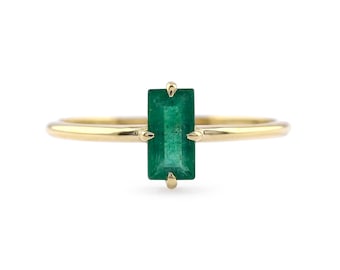May include: A gold ring featuring a single rectangular emerald gemstone set vertically in a four-prong setting. The band is thin, smooth, and polished, showcasing a minimalist design that highlights the deep green color of the central stone.