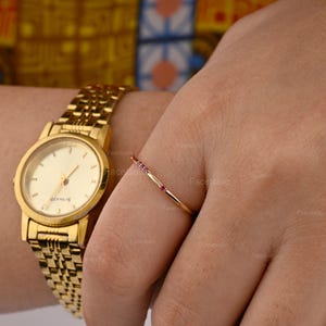 May include: A gold-toned analogue wristwatch with a metallic link strap and a round face featuring thin hands and markers, paired with a delicate gold ring set with four small, evenly spaced pink gemstones on a thin, polished gold band.