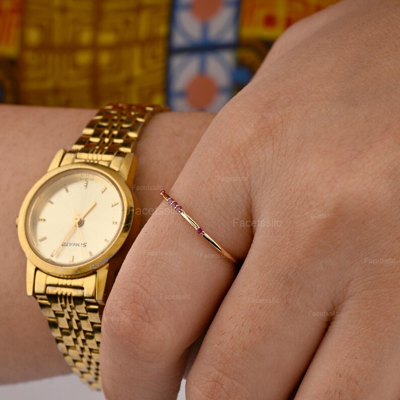 May include: A gold-toned analogue wristwatch with a metallic link strap and a round face featuring thin hands and markers, paired with a delicate gold ring set with four small, evenly spaced pink gemstones on a thin, polished gold band.