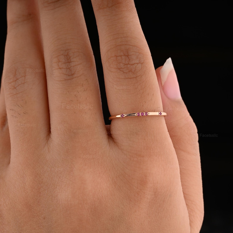 May include: A thin rose gold band ring featuring three small, round, pink gemstones set horizontally in the centre. The metal has a polished finish, and the design is simple and slender, showcasing a delicate appearance on the finger.