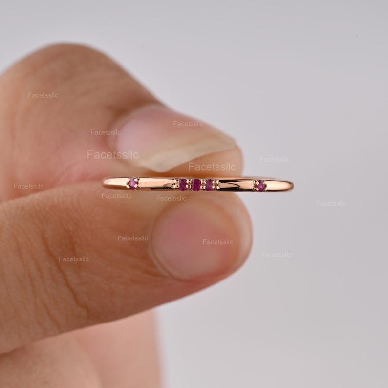 May include: A thin rose gold band ring featuring a horizontal row of five small, round, deep pink rubies set into the metal. The ring has a smooth, polished finish and a minimalist design, showcasing the vibrant red gemstones against the metal.
