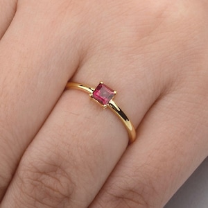 May include: A gold ring featuring a single, small, square-cut, deep pink gemstone set in a four-prong setting. The band is thin, smooth, and polished, encircling the finger with a minimalist design that highlights the vibrant color of the stone.