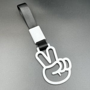 May include: A white plastic hand-shaped handle displaying a peace sign gesture, attached to a black nylon strap with a white rectangular connector. The handle features a hollow, stylized design and is oriented vertically against a dark background.