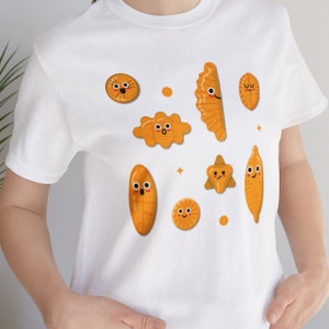 Könnte beinhalten: Weißes T-Shirt mit einem Cartoon-Design aus verschiedenen Gebäckstücken mit lächelnden Gesichtern. Das Gebäck ist in orange und gelben Farben gehalten.