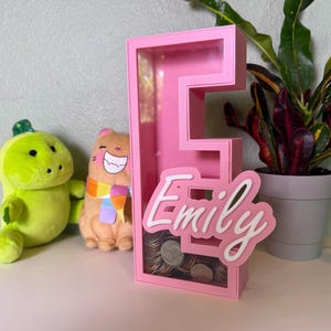 Può includere: Una salvadanaio rosa a forma di lettera "E" con il nome "Emily" in corsivo bianco. La banca contiene monete. Due peluche e una pianta in vaso sono sullo sfondo.