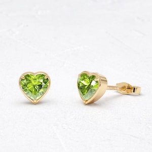 May include: A pair of gold stud earrings featuring heart-shaped, light green gemstones. The earrings are set in a gold bezel setting and have a post and butterfly clasp. The gemstones have a faceted cut.