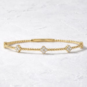 Może przedstawiać: Złota bransoletka z motywem koniczyny. Bransoletka posiada małe, okrągłe koraliki i trzy zawieszki w kształcie koniczyny, każda z wieloma małymi, przezroczystymi kamieniami. Bransoletka na białym tle.