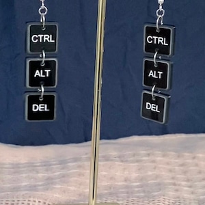 May include: A pair of black and white earrings with the words "CTRL", "ALT", and "DEL" on them. The earrings are hanging from a gold stand.