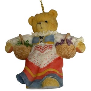 Op de afbeelding: Een gele teddybeer die een rood, wit en blauw jurkje draagt en twee manden met druiven draagt. De beer is een kerstversiering.