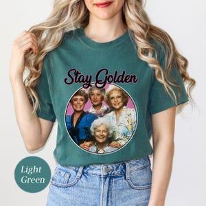 Op de afbeelding: Een lichtgroen T-shirt met een roze cirkel met een foto van vier vrouwen. De tekst "Stay Golden" staat boven de cirkel.