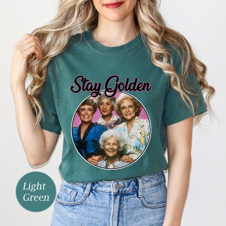 Op de afbeelding: Een lichtgroen T-shirt met een roze cirkel met een foto van vier vrouwen. De tekst "Stay Golden" staat boven de cirkel.