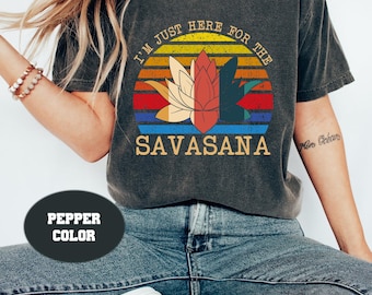 May include: A dark grey t-shirt with a vintage sunset graphic and the text "I'm just here for the Savasana". The graphic features a lotus flower in the centre of the sunset.