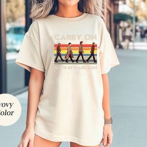 May include: A cream colored t-shirt with a retro graphic of four figures walking across a rainbow stripe. The text "Carry On My Wayward Son" is printed below the figures.