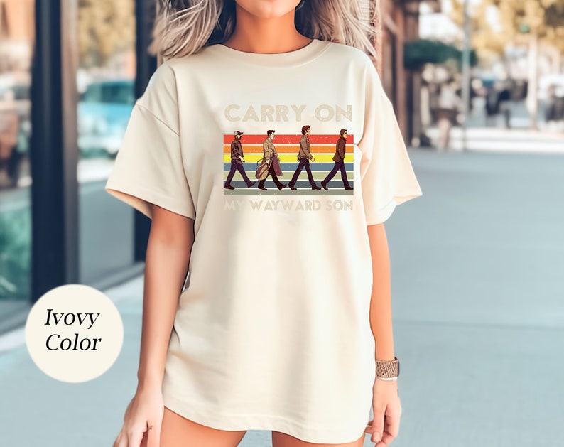 May include: A cream colored t-shirt with a retro graphic of four figures walking across a rainbow stripe. The text "Carry On My Wayward Son" is printed below the figures.