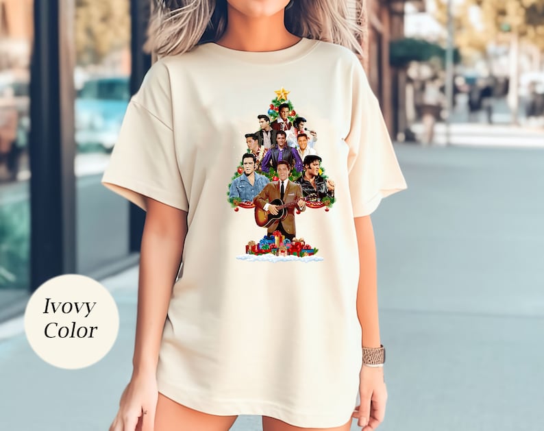 May include: A white t-shirt with a Christmas tree design featuring a collage of Elvis Presley images. The tree is decorated with ornaments and presents. The text "Elvis Christmas" is at the top of the tree.