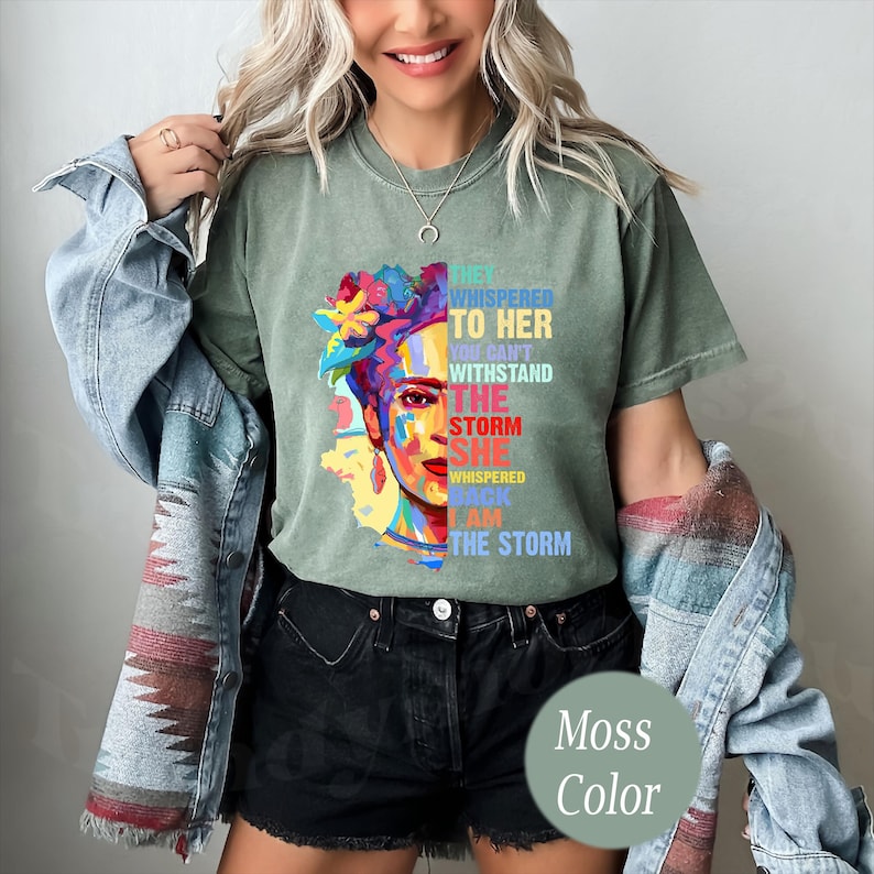 Pu&ograve; includere: T-shirt verde muschio con un ritratto colorato di una donna con decorazioni floreali. Il testo sulla maglietta recita "They whispered to her... I am the storm." La maglietta &egrave; di vestibilit&agrave; ampia.