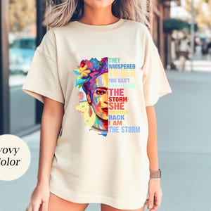 Pu&ograve; includere: T-shirt color avorio con un ritratto colorato di una donna con copricapo floreale. Il testo sulla maglietta recita: "Le sussurrarono che non puoi resistere alla tempesta, lei sussurr&ograve; che io sono la tempesta."