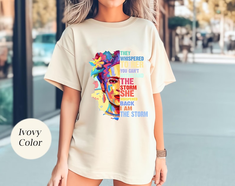 Pu&ograve; includere: T-shirt color avorio con un ritratto colorato di una donna con copricapo floreale. Il testo sulla maglietta recita: "Le sussurrarono che non puoi resistere alla tempesta, lei sussurr&ograve; che io sono la tempesta."