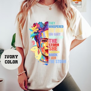 Pu&ograve; includere: T-shirt color avorio con una grafica colorata del volto di una donna con accenti floreali e il testo: "Le sussurravano, non puoi resistere alla tempesta. Lei sussurr&ograve;, io sono la tempesta."