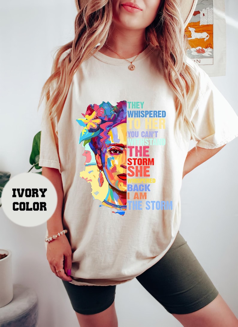 Pu&ograve; includere: T-shirt color avorio con una grafica colorata del volto di una donna con accenti floreali e il testo: "Le sussurravano, non puoi resistere alla tempesta. Lei sussurr&ograve;, io sono la tempesta."
