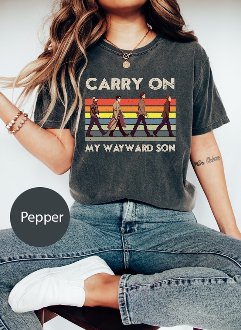 May include: A dark gray t-shirt with a vintage-style graphic of four figures walking across a rainbow stripe background. The text "Carry On My Wayward Son" is printed below the graphic.