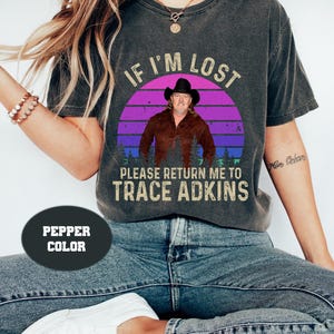 May include: A dark grey t-shirt with a vintage-style graphic of a man in a cowboy hat. The graphic says "If I'm Lost Please Return Me To Trace Adkins".