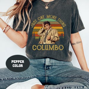 May include: A vintage-style graphic tee shirt with a faded black background featuring a retro sunset graphic with the text "Just One More Thing" and the name "Columbo" underneath. The graphic features a man in a trench coat with a worried expression.