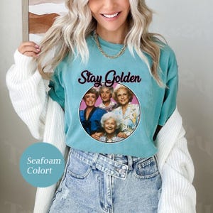 Op de afbeelding: Een turquoise t-shirt met een roze cirkelvormige afbeelding met vier vrouwen en de tekst "Stay Golden".