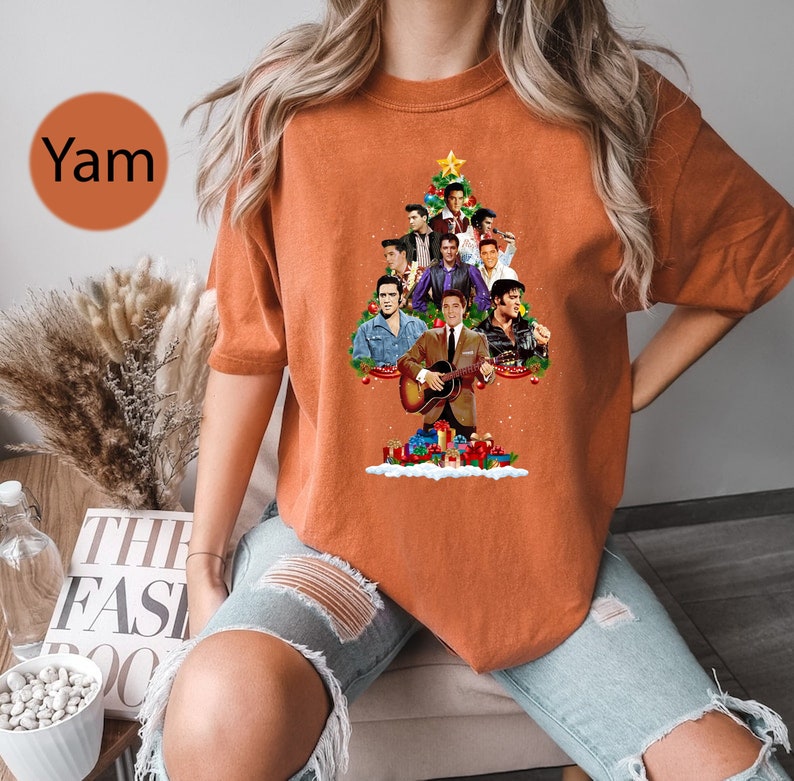 May include: Orange t-shirt with a Christmas tree design featuring multiple images of Elvis Presley. The tree is decorated with ornaments and presents.