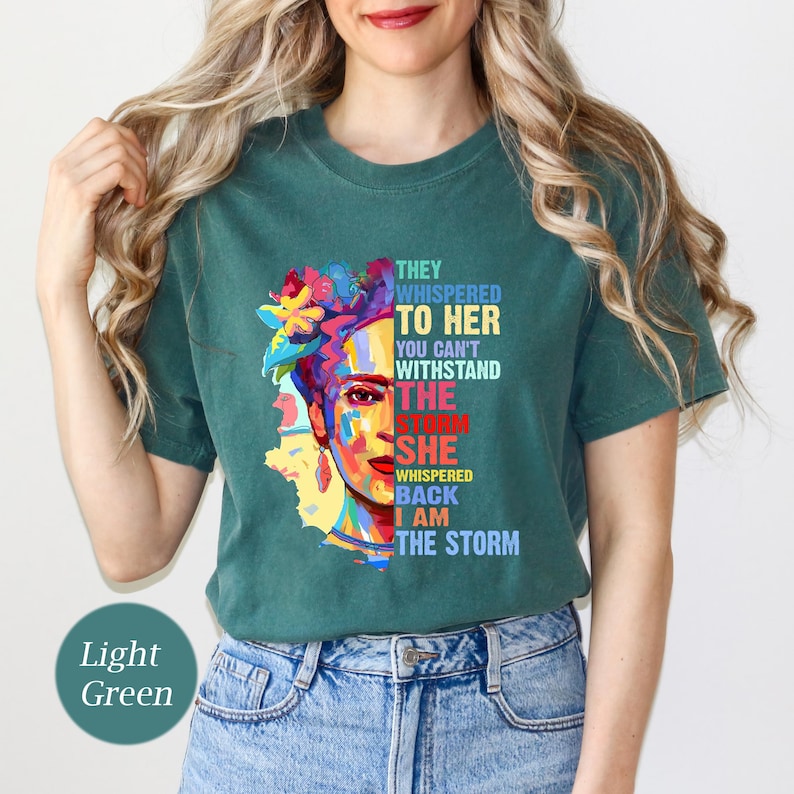 Pu&ograve; includere: T-shirt verde chiaro con una grafica colorata del volto di una donna con accenti floreali. Il testo recita: "Le sussurravano, non puoi resistere alla tempesta. Lei sussurr&ograve;, io sono la tempesta."
