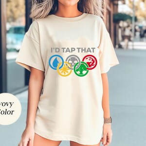 Pode incluir: Uma camiseta cor creme com o texto "I'd Tap That" e um gr&aacute;fico de cinco an&eacute;is ol&iacute;mpicos com s&iacute;mbolos de Magic: The Gathering.