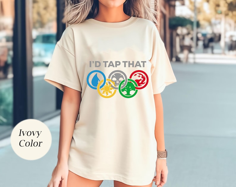 Pode incluir: Uma camiseta cor creme com o texto "I'd Tap That" e um gr&aacute;fico de cinco an&eacute;is ol&iacute;mpicos com s&iacute;mbolos de Magic: The Gathering.