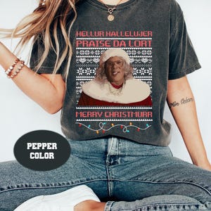 Könnte beinhalten: Dunkelgraues T-Shirt mit einem Weihnachtspullover-Design und dem Schriftzug "HELLUR HALLELUJER PRAISE DA LORT MERRY CHRISTMURR". Das Design zeigt ein Porträt einer Figur in einem weihnachtlichen Outfit und eine Lichterkette.