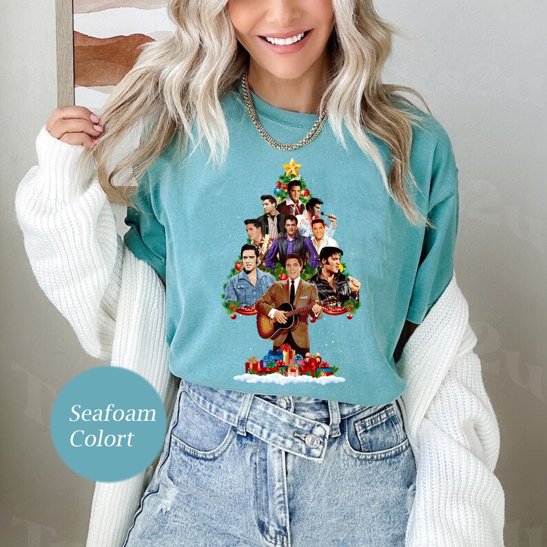 May include: A teal blue t-shirt with a Christmas tree design featuring a collage of images of Elvis Presley. The tree is decorated with red and green ornaments and a star on top.