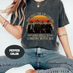 May include: A dark grey t-shirt with a vintage-style graphic of four men in cowboy hats walking across a sunset with the text "You tell 'em I'm coming and hell's coming with me".