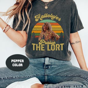 May include: A dark gray t-shirt with a vintage-style sunset graphic featuring a woman with the text "Hallelujah Praise The Lort".
