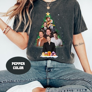 Peut inclure: T-shirt gris foncé avec un motif de sapin de Noël composé d'images d'un homme. L'arbre est surmonté d'une étoile dorée et entouré de flocons de neige. Le t-shirt porte également le texte "PEPPER COLOR" dans un ovale foncé.