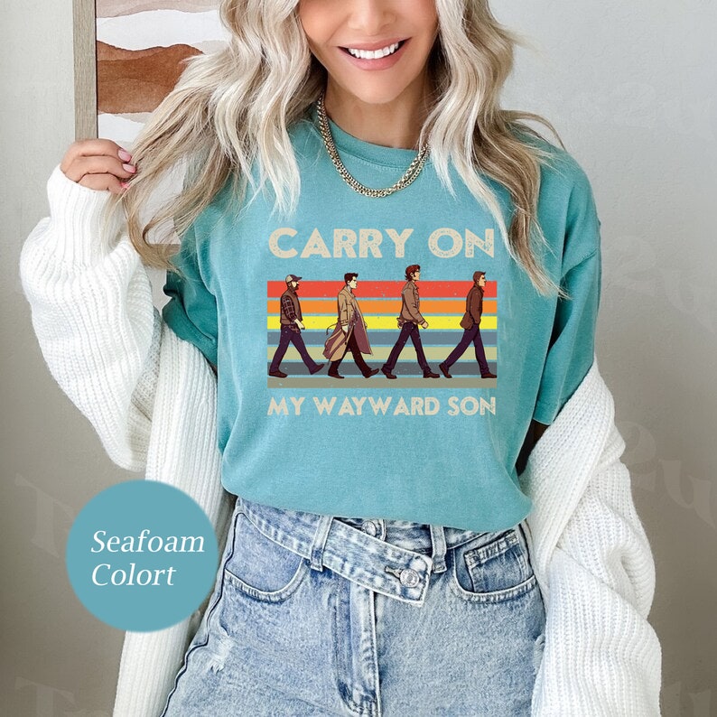 May include: A teal blue t-shirt with a vintage-style graphic of four figures walking across a rainbow striped background. The text "Carry On My Wayward Son" is printed below the figures.