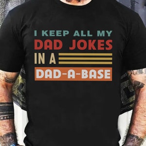 May include: Black t-shirt with a vintage-style graphic that reads "I keep all my dad jokes in a dad-a-base".