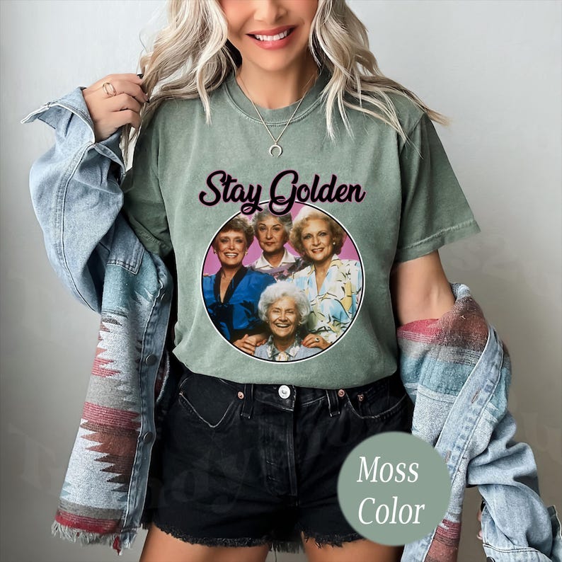 Op de afbeelding: Een groen T-shirt met een vintage look met een afbeelding van de Golden Girls met de tekst "Stay Golden" erop gedrukt. Het shirt wordt gedragen door iemand die een zwarte spijkerbroek en een blauw spijkerjack draagt.