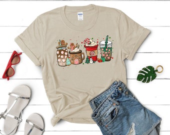 Op de afbeelding: Een beige T-shirt met een afbeelding van vier feestelijke drankjes. De drankjes zijn een gingerbread latte, een peppermint mocha, warme chocolademelk en een peppermint mocha iced coffee. De afbeelding is in cartoon-stijl en heeft feestelijke kleuren zoals rood, groen en wit.