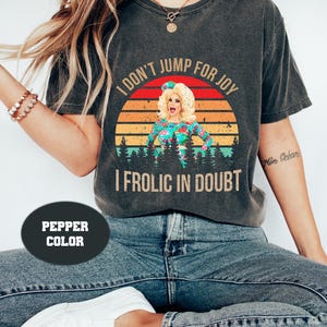 May include: A dark grey t-shirt with a vintage-style sunset graphic and the text "I don't jump for joy I frolic in doubt". The graphic features a woman with blonde hair and a floral dress.