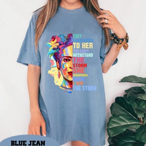 Pu&ograve; includere: Una maglietta blu con una grafica colorata del volto di una donna con accenti floreali e la scritta "THEY WHISPERED TO HER YOU CAN'T WITHSTAND THE STORM SHE WHISPERED I AM THE STORM". La maglietta ha maniche corte.